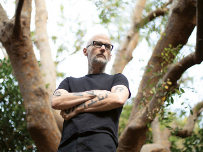 Moby standing with arms folded in front of a tree